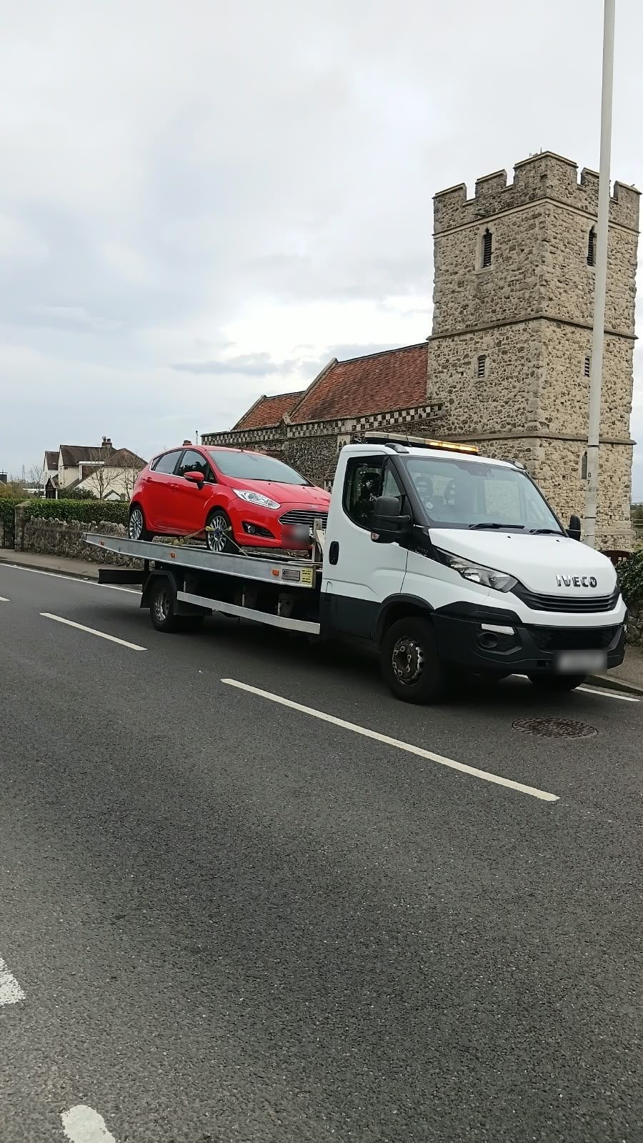Towing service truck carrying a red car past a historic church tower